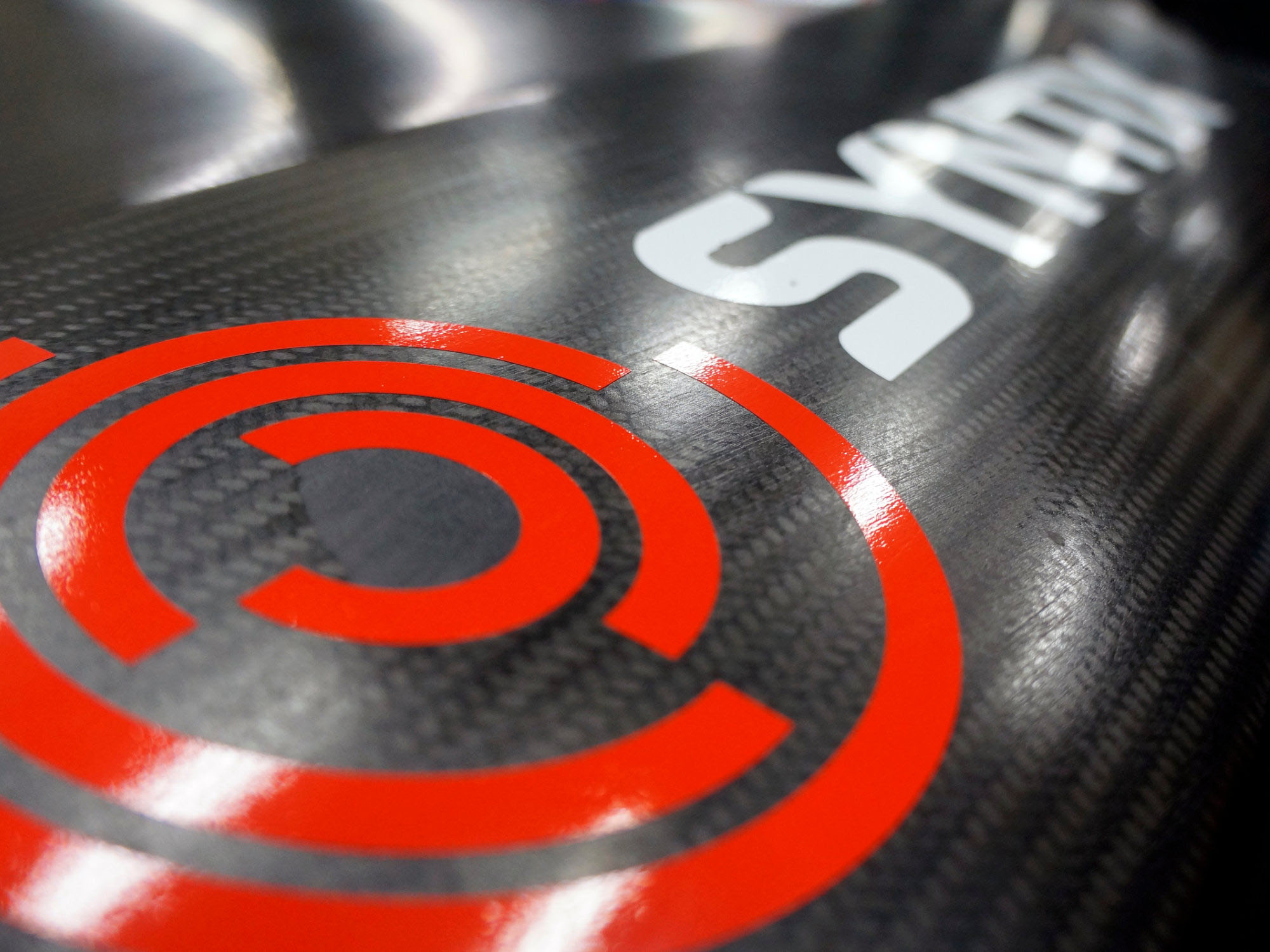 Close-Up of Syntix Logo on Carbon Fiber Surface Close-up view of the Syntix logo with red concentric icon on a carbon fiber background