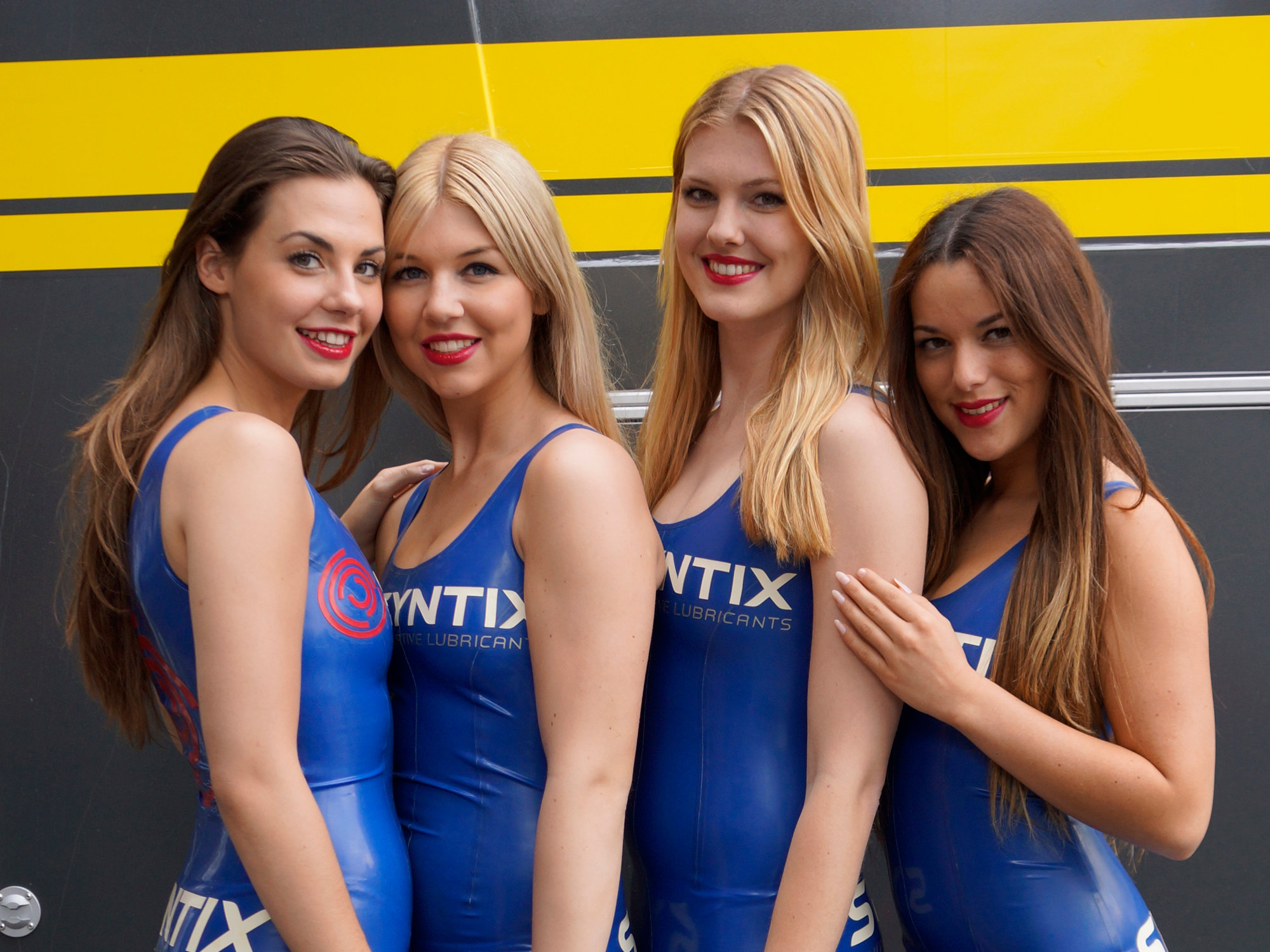 Grid girls wearing red and blue Syntix-branded latex suits at a racing event